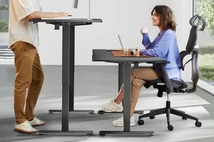 A woman sits at a standing desk using a laptop, while a man stands at a similar desk in a modern office.