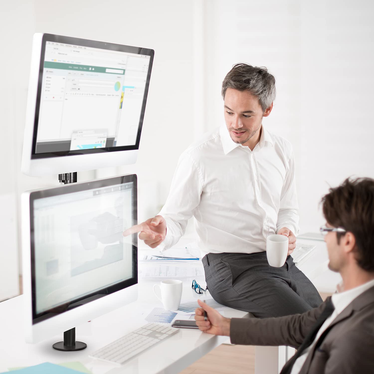 Two professionals discussing work on a vertical dual monitor setup in an office.